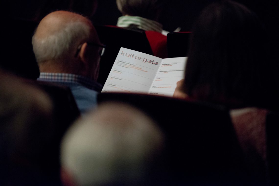 Das Bild zeigt die Rückansicht eines Gastes, welcher im Stadttheater sitzt und im Programm der Kulturgala liest.
