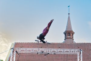 Das Foto zeigt eine Performance des Bielefelder Sommertheaters. Hierbei fliegt eine Artistin durch die Luft, während sie sich an einem Trapez festhält.